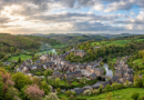 A quaint village with stone houses, a church with a tall steeple, a castle on a hill, and a river winding through green countryside
